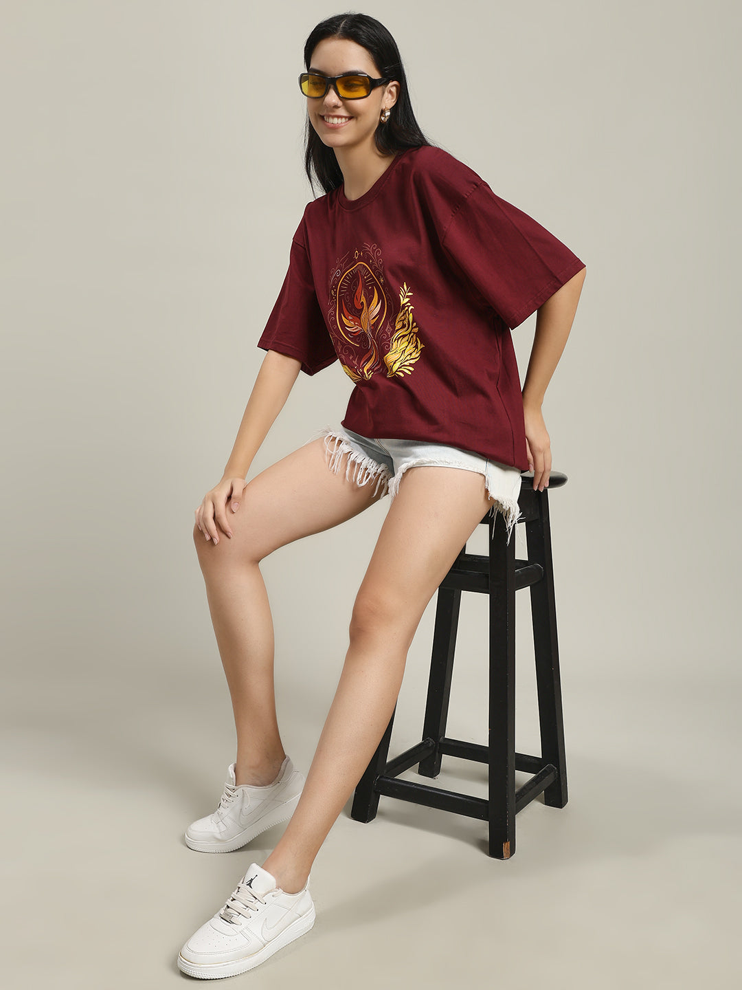 Woman sitting on a black stool wearing a maroon Fire women's over-sized T-shirt with phoenix and fire design, paired with light denim shorts and white sneakers, adjusting yellow-tinted sunglasses.
