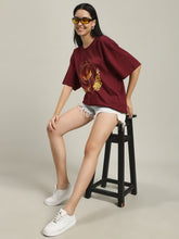 Woman sitting on a black stool wearing a maroon Fire women's over-sized T-shirt with phoenix and fire design, paired with light denim shorts and white sneakers, adjusting yellow-tinted sunglasses.