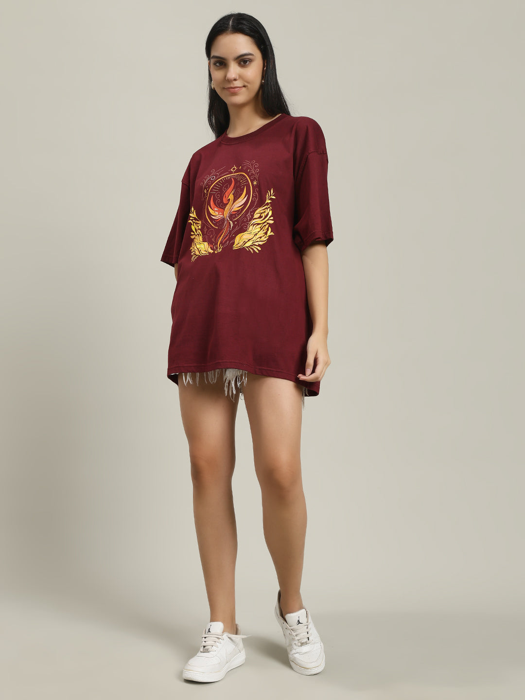 Front view of a woman smiling, wearing a maroon Fire women's over-sized T-shirt featuring a yellow and orange phoenix and fire design, paired with distressed white denim shorts and white sneakers