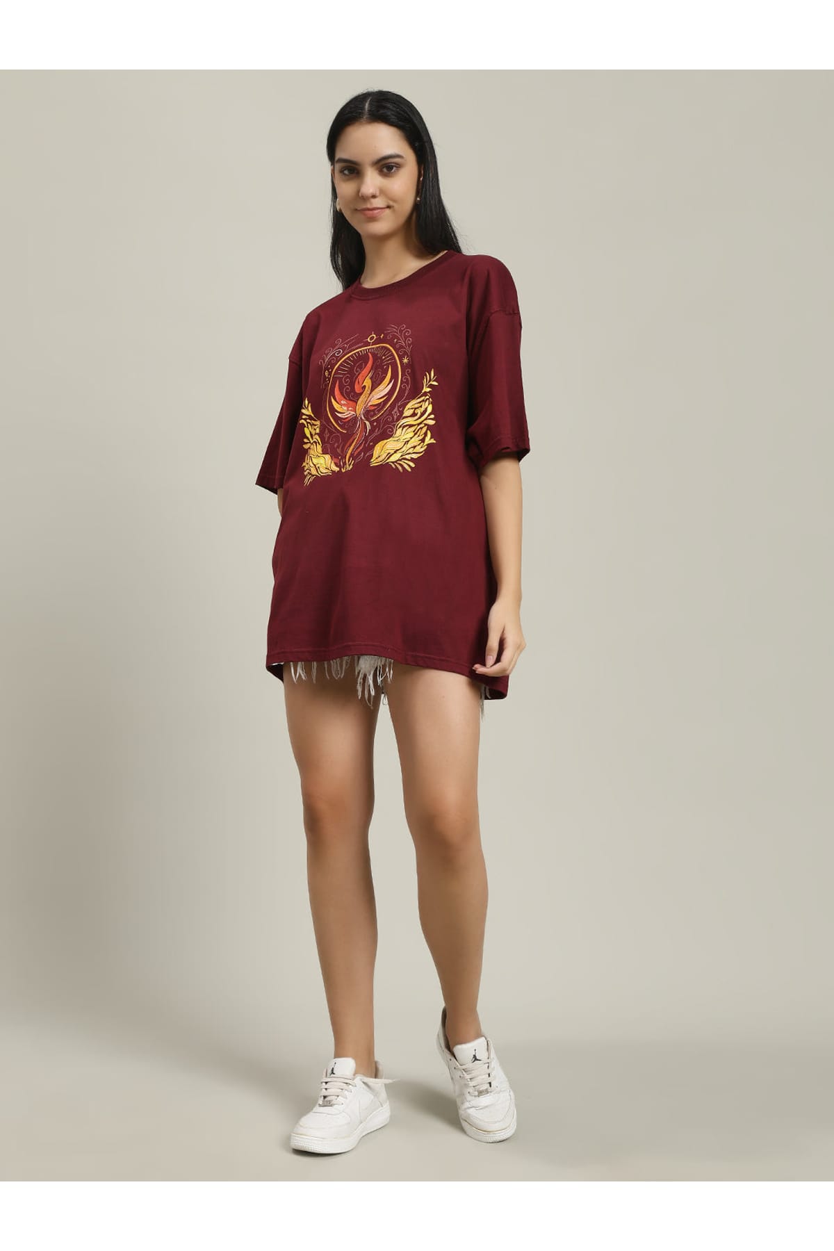 Front view of a woman smiling, wearing a maroon Fire women's over-sized T-shirt featuring a yellow and orange phoenix and fire design, paired with distressed white denim shorts and white sneakers