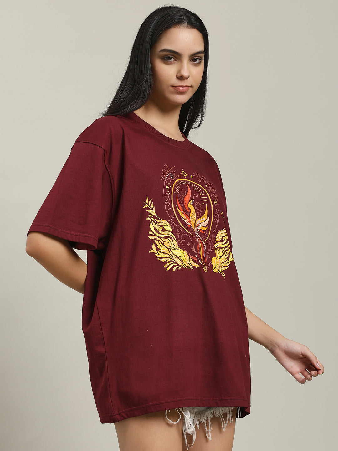 Side angle of a woman wearing the maroon Fire women's over-sized T-shirt with a phoenix fire graphic on the front, paired with distressed denim shorts, standing and smiling.