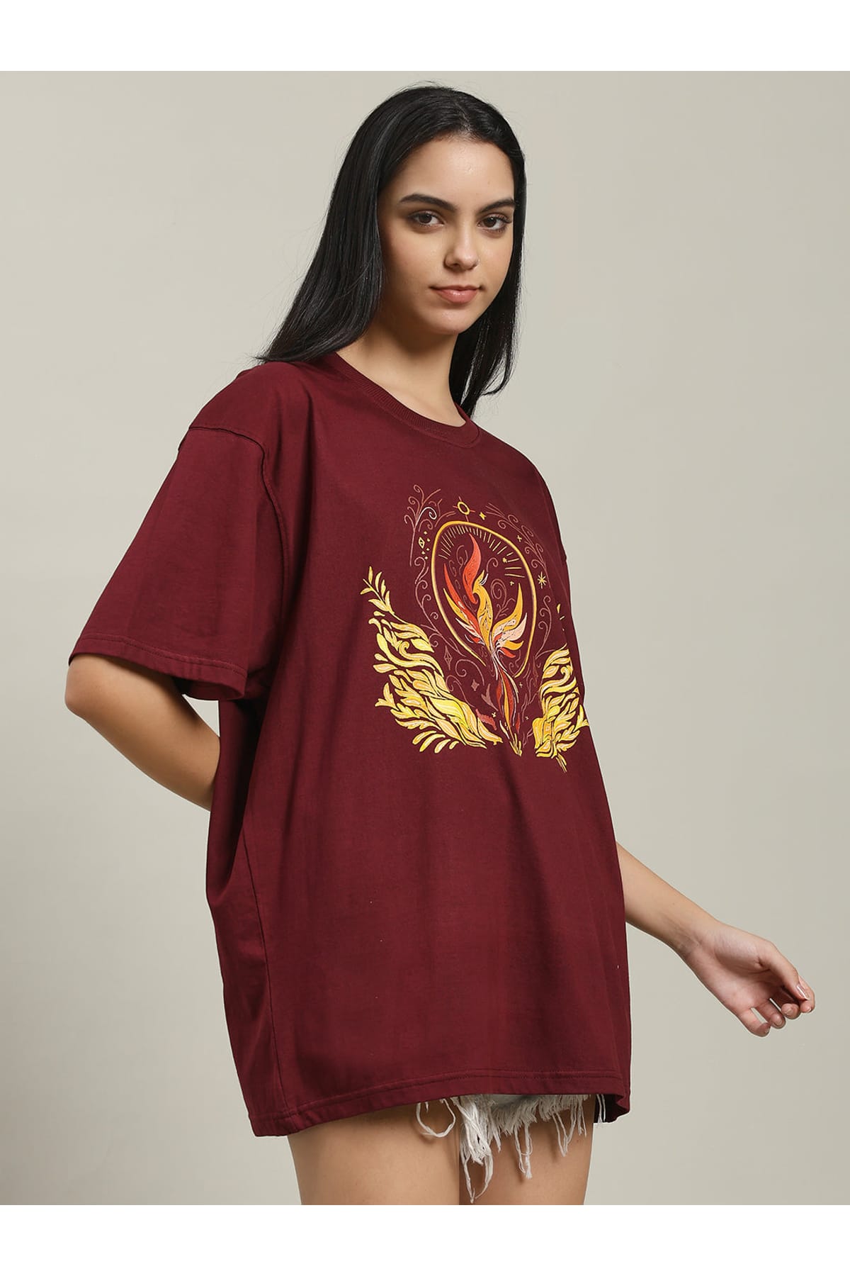 Side angle of a woman wearing the maroon Fire women's over-sized T-shirt with a phoenix fire graphic on the front, paired with distressed denim shorts, standing and smiling.