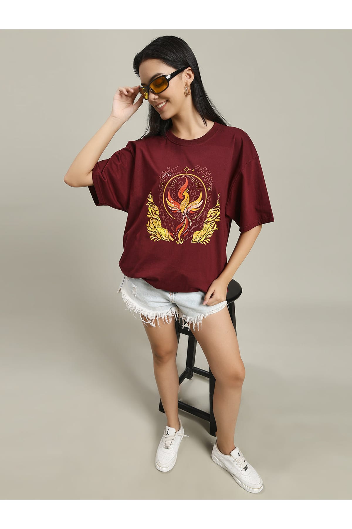 Woman standing and smiling, wearing a maroon Fire women's over-sized T-shirt with a phoenix fire print on the front, paired with white distressed shorts and white sneakers, posing against a neutral background