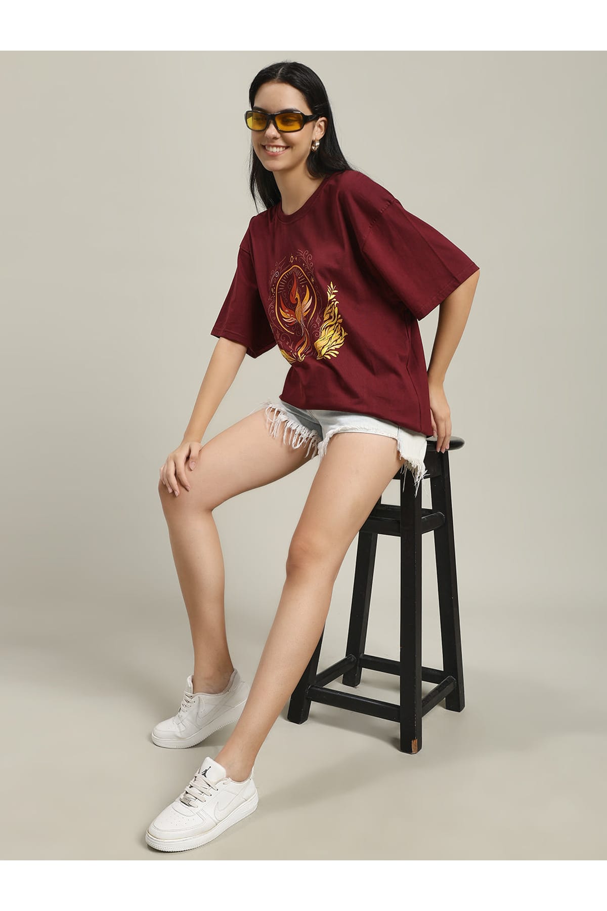 Woman sitting on a black stool wearing a maroon Fire women's over-sized T-shirt with phoenix and fire design, paired with light denim shorts and white sneakers, adjusting yellow-tinted sunglasses.